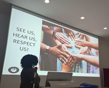 Bild von Esinu Afele vor einer Slideprojektion mit dem Appell "See us, hear us, respect us"