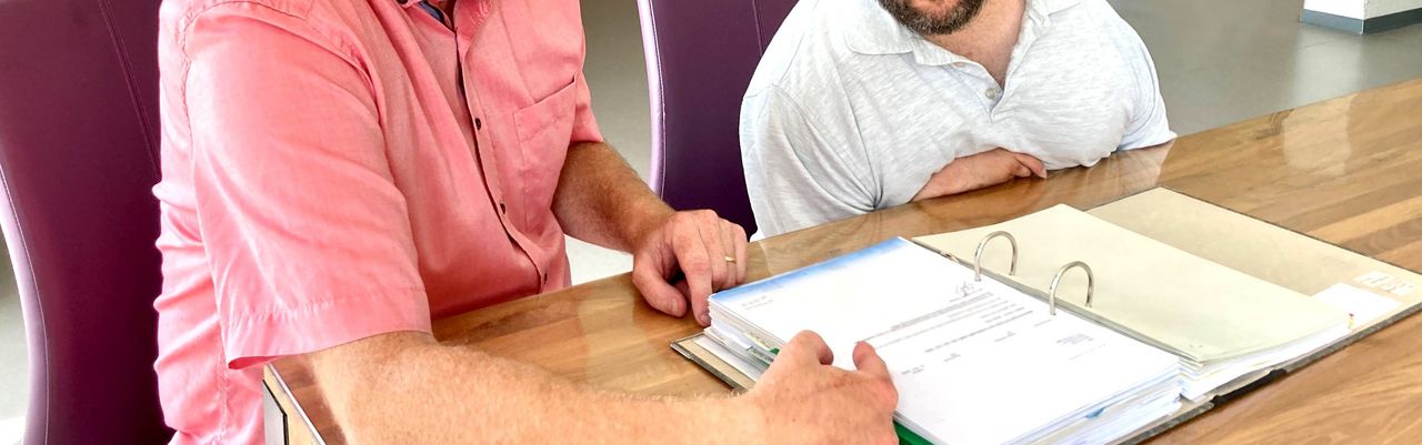 Ein rechtlicher Betreuer sitzt mit seinem Klienten am Tisch und zeigt ihm einen Brief in einem Ordner