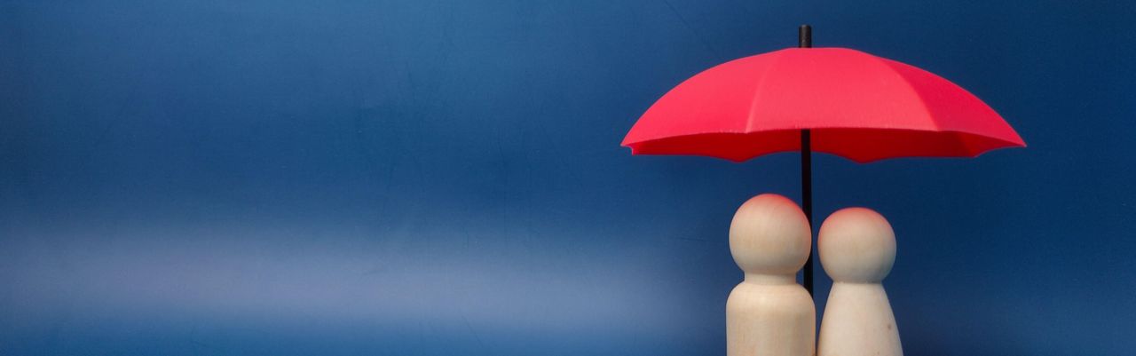 Zwei Holzfiguren stehen unter einem roten Schirm, der ihnen Schutz bietet